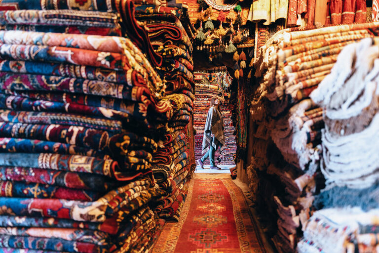 Woman browses stacks of fine rugs in a market.