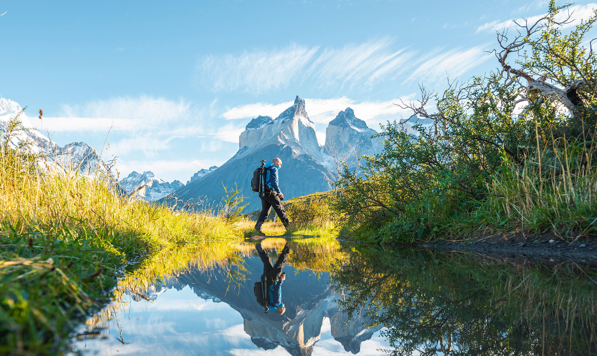 A hiker with photography equipment crosses a pond while sharp mountain peaks rise in the distance.