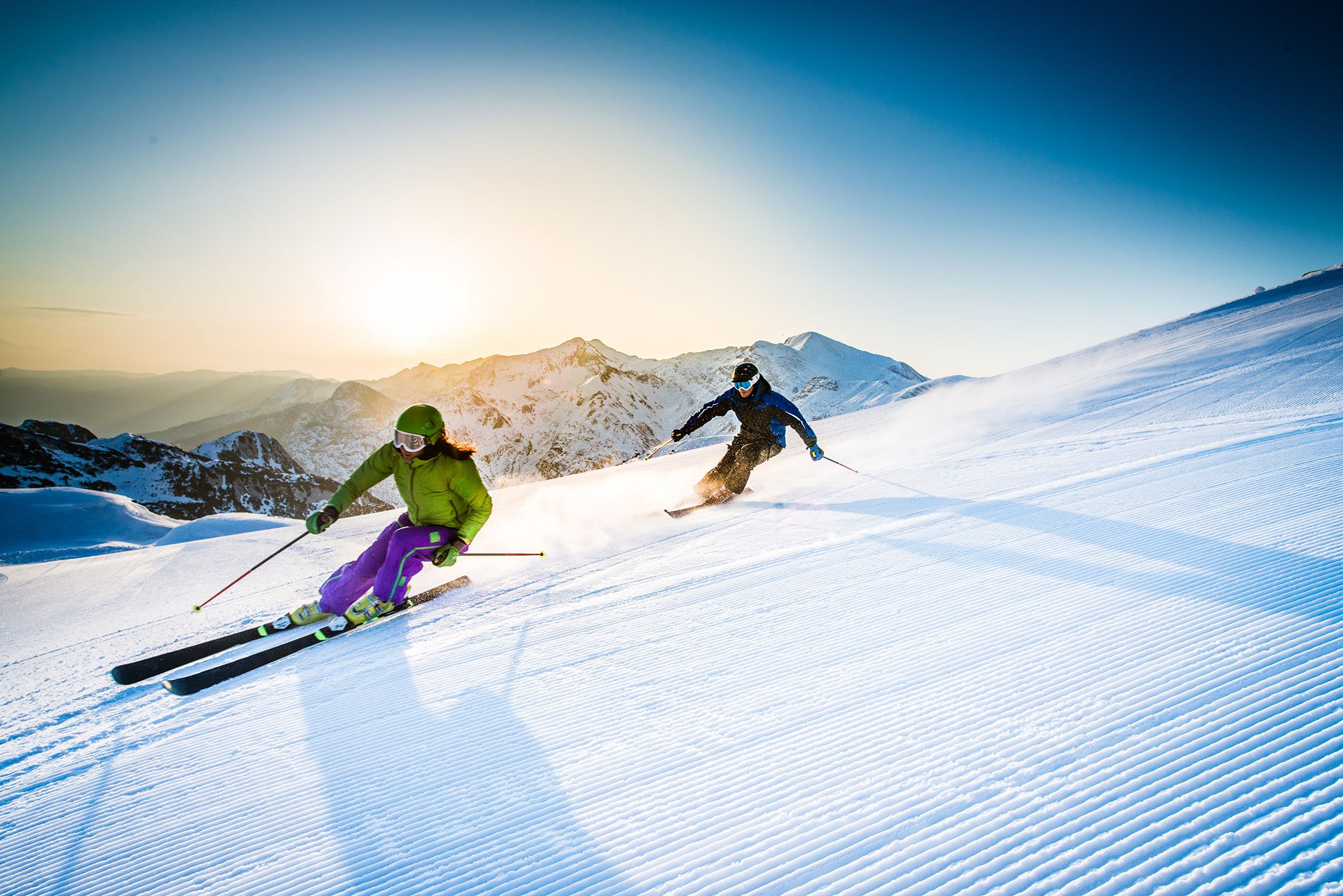Two snow skiers lean into a turn on fresh powder.